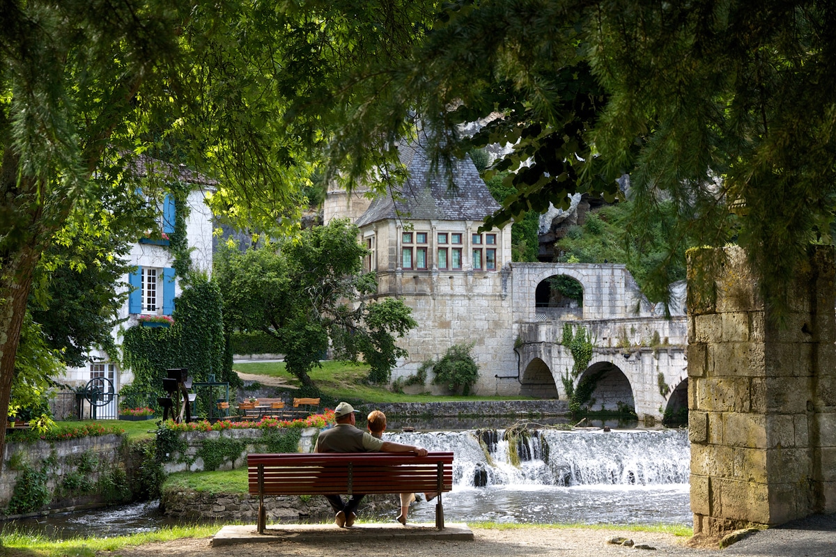 village de brantome avec un banc avec son père et son fils devant de l'eau et des maisons en fond