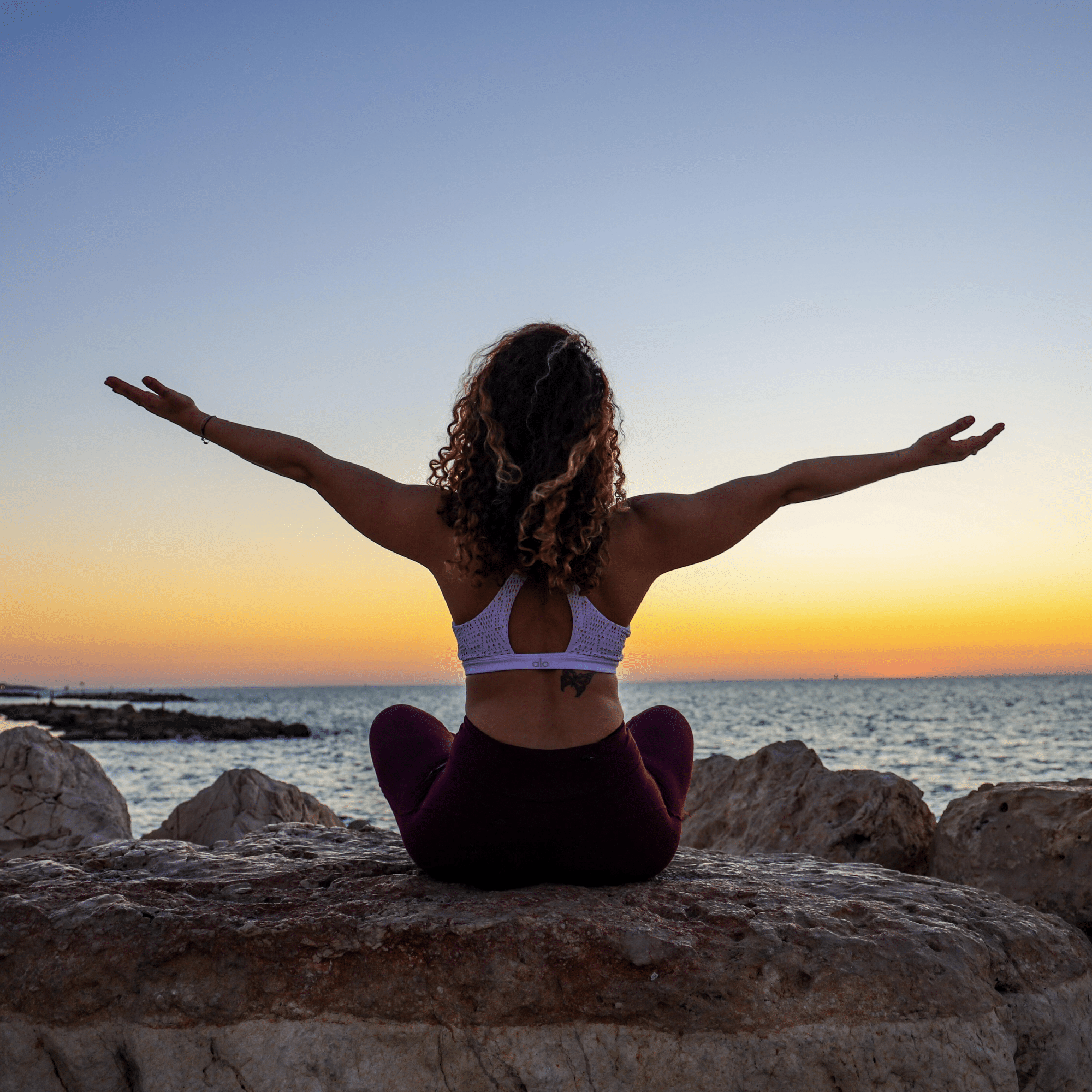 femme en tenue de sport assise face à la mer sur une pierre avec le couché du soleil